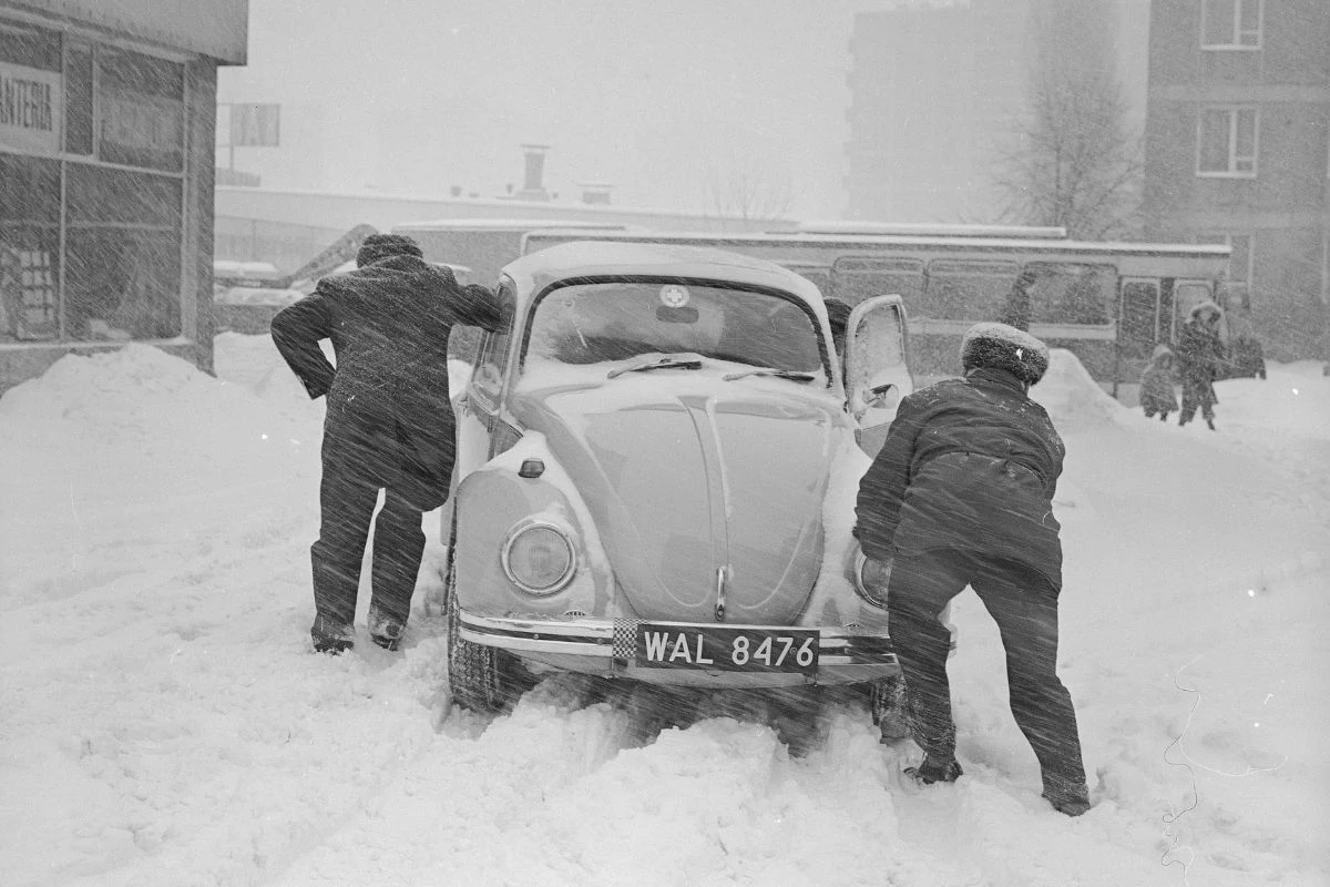 Zimą w Warszawie nie brakowało wyzwań Dwóch mężczyzn próbuje wypchnąć samochód Volkswagen Garbus z głębokiego śniegu w środku miasta podczas intensywnej śnieżycy. Za pojazdem widoczny jest charakterystyczny autobus, a w tle zarysowują się wysokie budynki mieszkalne oraz piesi zmagający się...