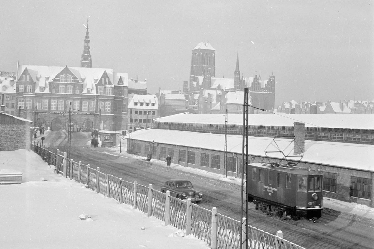 Tak wyglądał Gdańsk w 1959 roku Zimowy krajobraz miejskiej ulicy pokrytej śniegiem, po której jedzie niewielki tramwaj oraz samochód. W tle widać historyczne budynki z charakterystyczną wieżą i dachami pokrytymi śniegiem, spacerujących przechodniów i ogrodzenie oddzielające drogę od ...