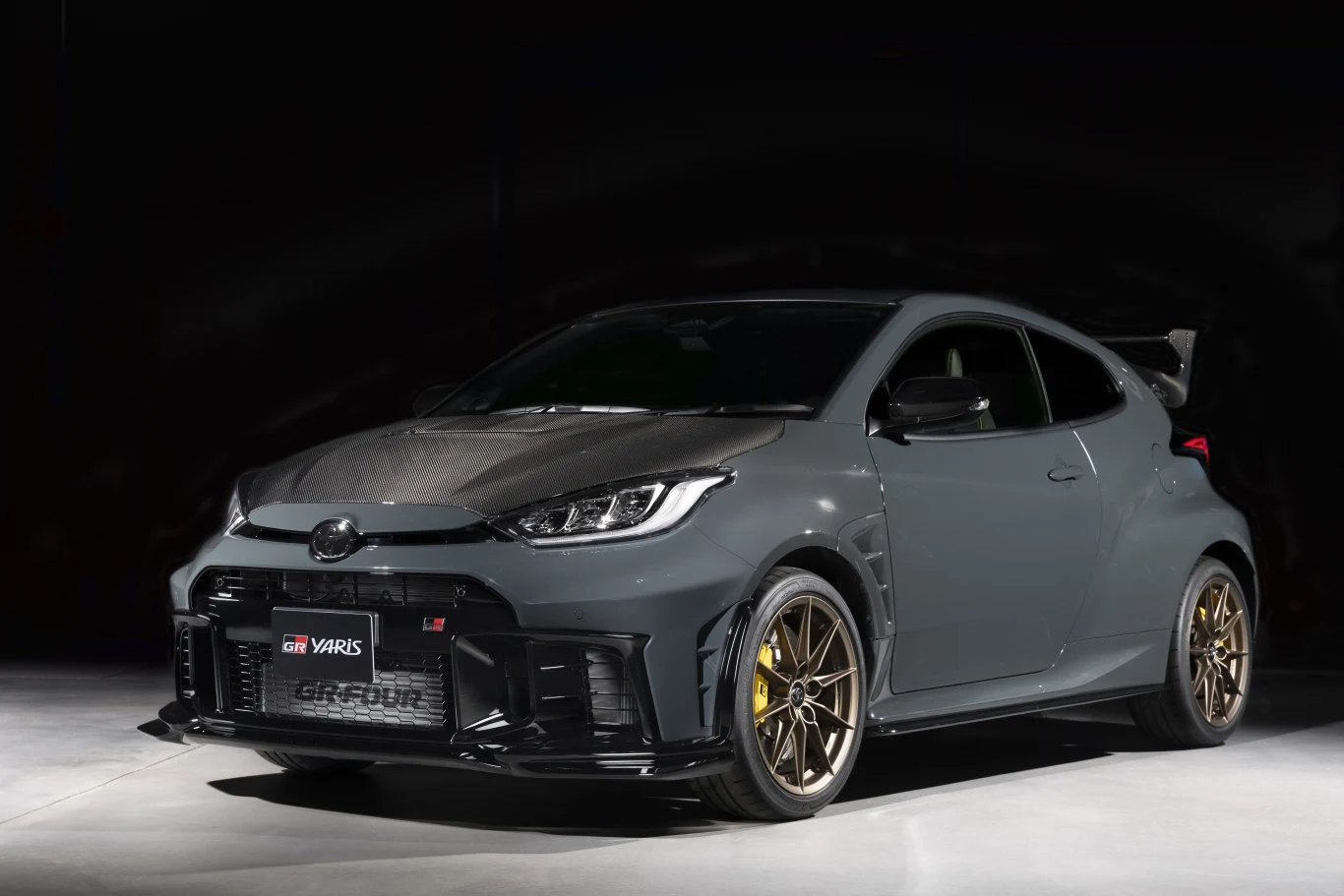 Szary sportowy samochód Toyota GR Yaris z charakterystyczną karbonową maską, sportowym pakietem stylistycznym, dużym tylnym spojlerem i złotymi felgami, ustawiony na tle ciemnego, jednolitego tła.