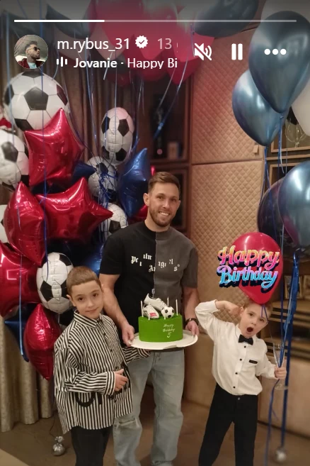 Mężczyzna trzyma tort urodzinowy ozdobiony motywami piłkarskimi, obok dwóch chłopców, jeden w eleganckiej koszuli i drugi pozujący z humorem. W tle balony z motywami piłki nożnej oraz napis Happy Birthday.