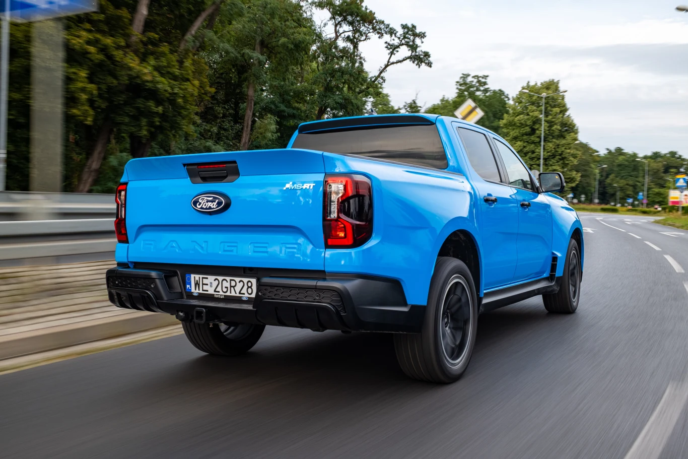 Niebieski pickup Ford Ranger Raptor jedzie asfaltową drogą przez zielony teren, mając czarne felgi i przyciemniane tylne szyby, a w tle widać drzewa oraz barierki ochronne.