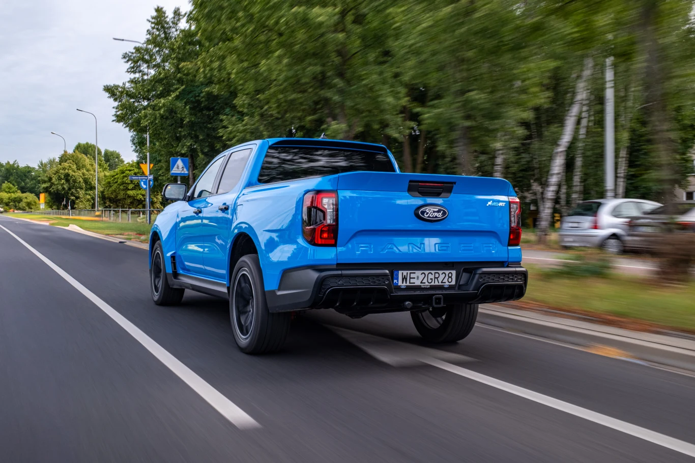 Niebieski pickup Ford Ranger porusza się po dwupasmowej, asfaltowej drodze otoczonej zielonymi drzewami; w tle znaki drogowe i fragmenty chodnika.