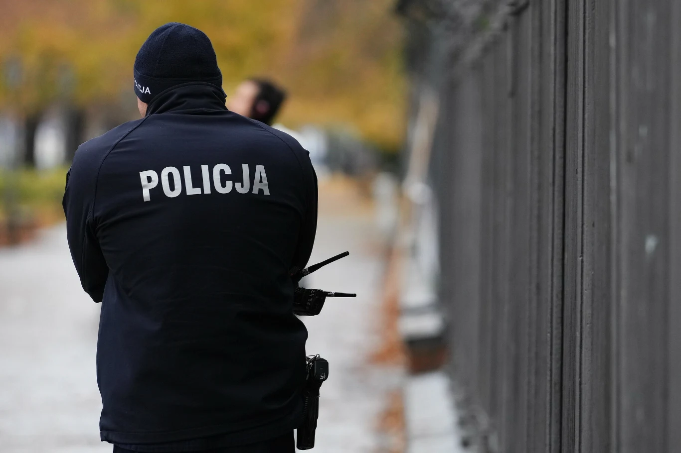 28-latka twierdzi, że molestował ją policjant (zdj. ilustracyjne) Funkcjonariusz policji w mundurze stoi tyłem przy ogrodzeniu, w tle widoczna rozmyta postać oraz jesienne drzewa.