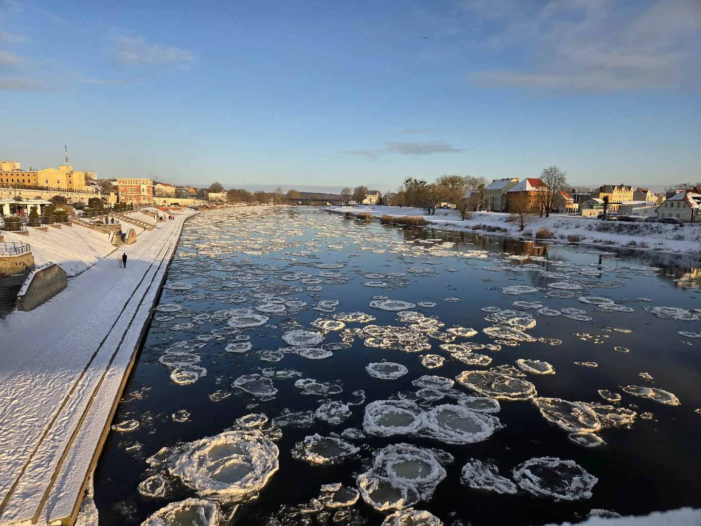 Zamarznięte krę lodowe unoszące się na szerokiej rzece, brzeg pokryty śniegiem, nieliczne osoby spacerujące wzdłuż nabrzeża, zabudowania miejskie w tle oświetlone popołudniowym słońcem.