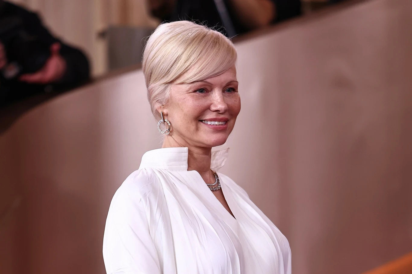 Pamela Anderson w ciągu ostatnich miesięcy eksperymentowała z kolorem włosów Kobieta o jasnych blond włosach upiętych w elegancki kok, ubrana w białą koszulę z szerokim kołnierzem, delikatny makijaż, uśmiecha się subtelnie, nosi duże okrągłe kolczyki oraz naszyjnik, tło rozmyte w odcieniach beżu.