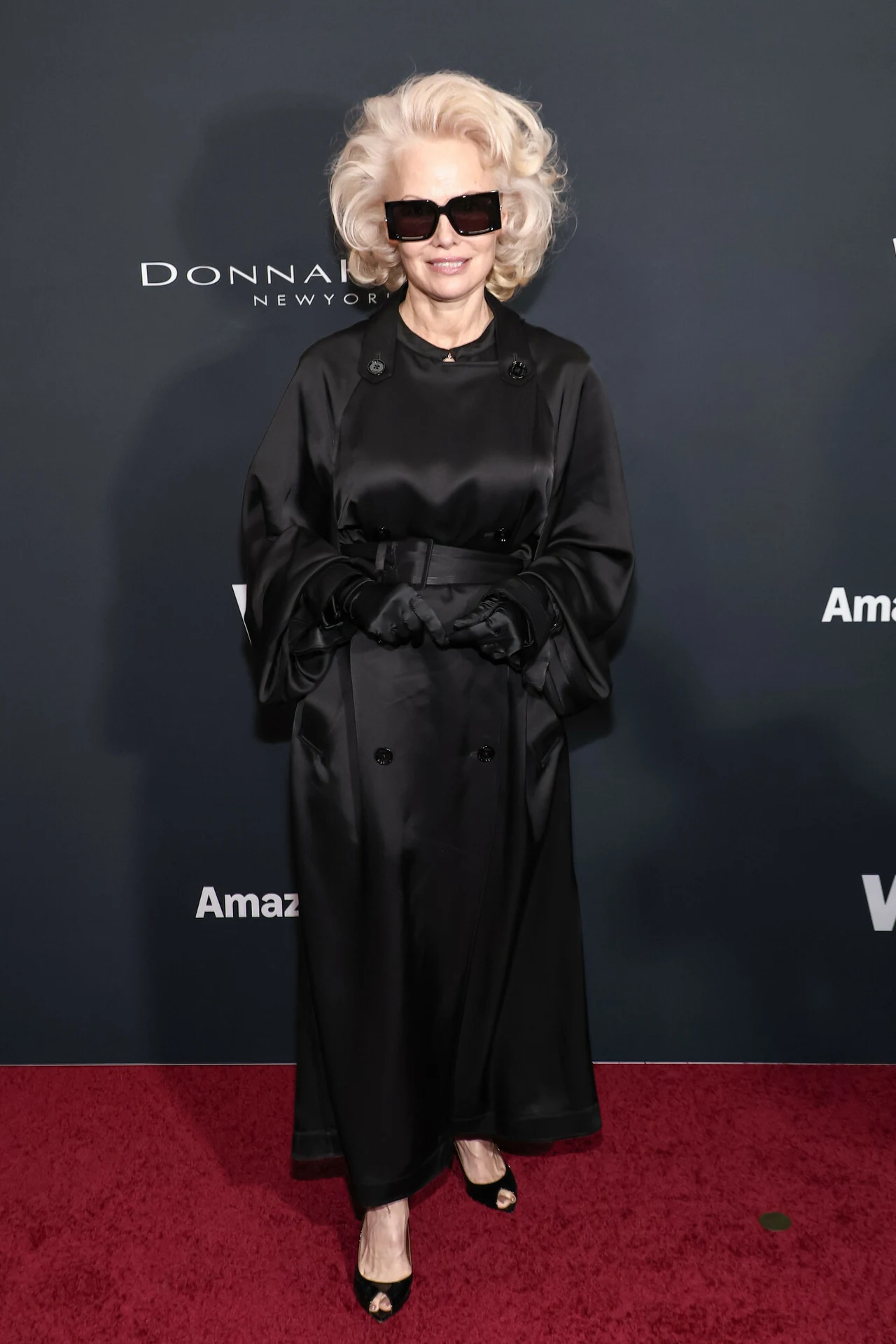 Pamela Anderson podczas Women’s Wear Daily Awards WWD Style Awards Elegancka kobieta w czarnej, długiej sukni i rękawiczkach, z rozpuszczonym blond włosami, dużymi okularami przeciwsłonecznymi i czarnymi butami na obcasie stoi na czerwonym dywanie na tle ściany z logotypami.