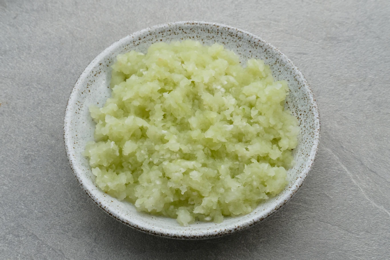 Talerz wypełniony drobno posiekaną lub startą masą o jasnozielonym kolorze, ułożoną na szarej powierzchni.