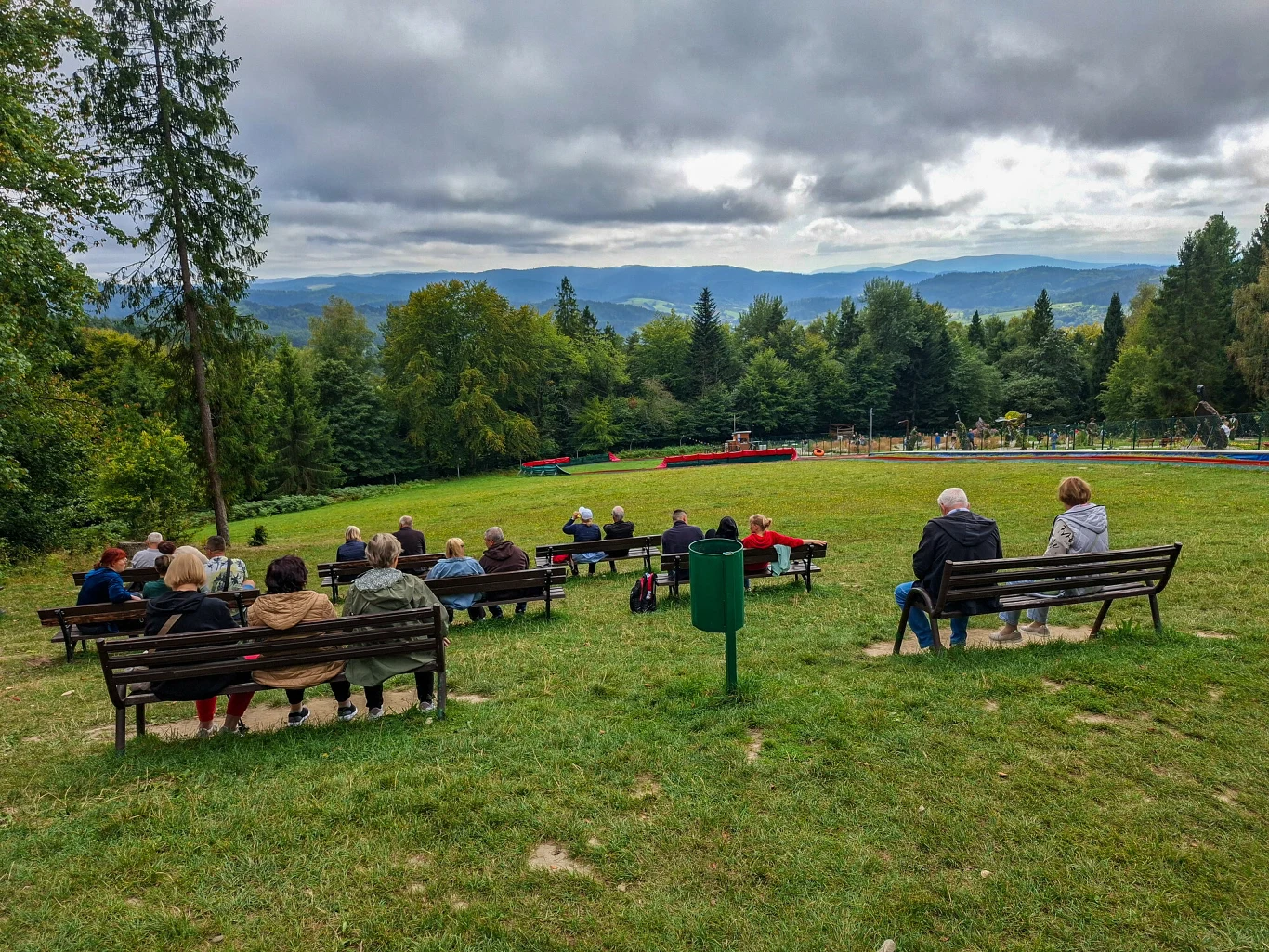 Widok ze szczytu Góry Parkowej jest piękny, ale ograniczony do jednego kierunku i przysłonięty przez drzewa Grupa ludzi siedzących na ławkach na zielonej łące, w otoczeniu drzew, z widokiem na góry w tle pod pochmurnym niebem.