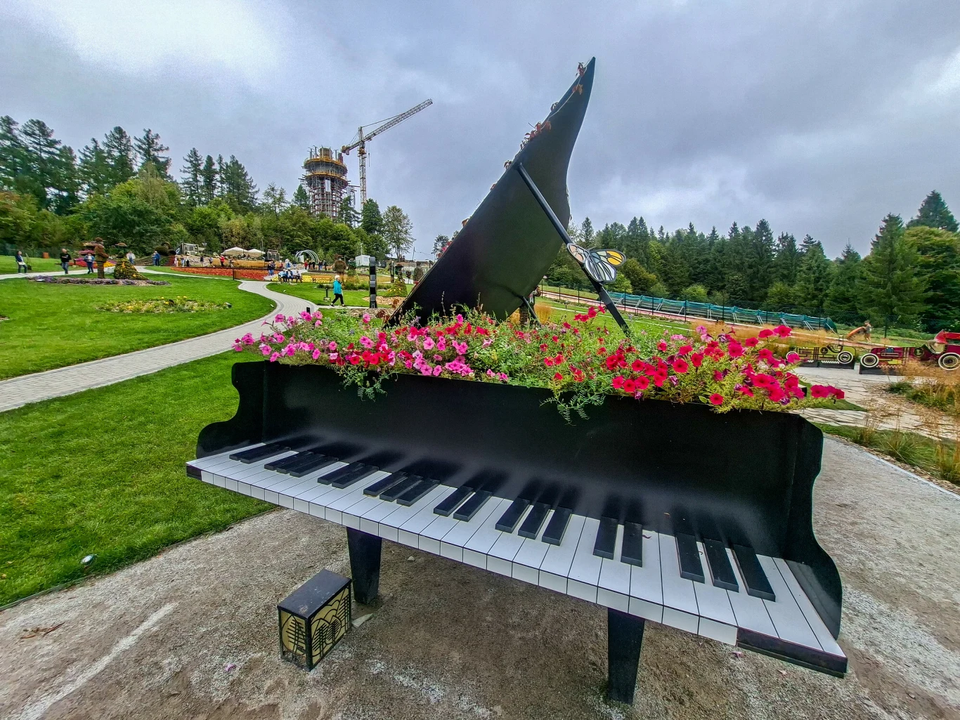 Polana Muzyki i Świateł na Górze Parkowej w Krynicy-Zdrój Czarny fortepian ogrodowy wypełniony kolorowymi kwiatami ustawiony na ścieżce w parku, w tle drzewa, zielona trawa, alejki oraz plac budowy z dźwigiem.