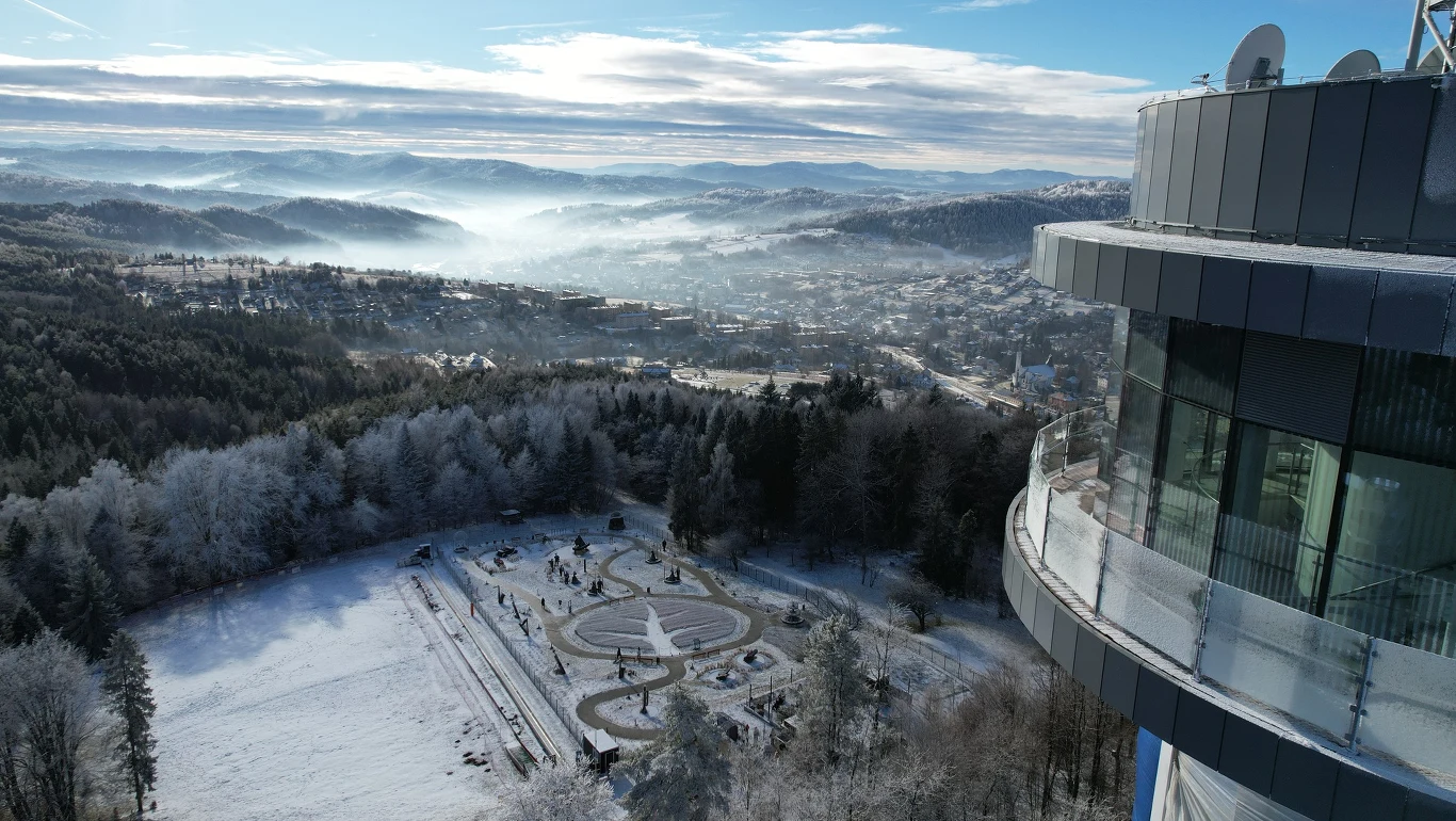 Nowa wieża widokowa w Krynicy-Zdroju gotowa na ferie zimowe Panorama zaśnieżonego parku z widoczną fontanną i alejkami, w tle rozległe pasmo gór oraz mgła unosząca się nad lasami, po prawej stronie nowoczesny budynek z przeszklonym tarasem.