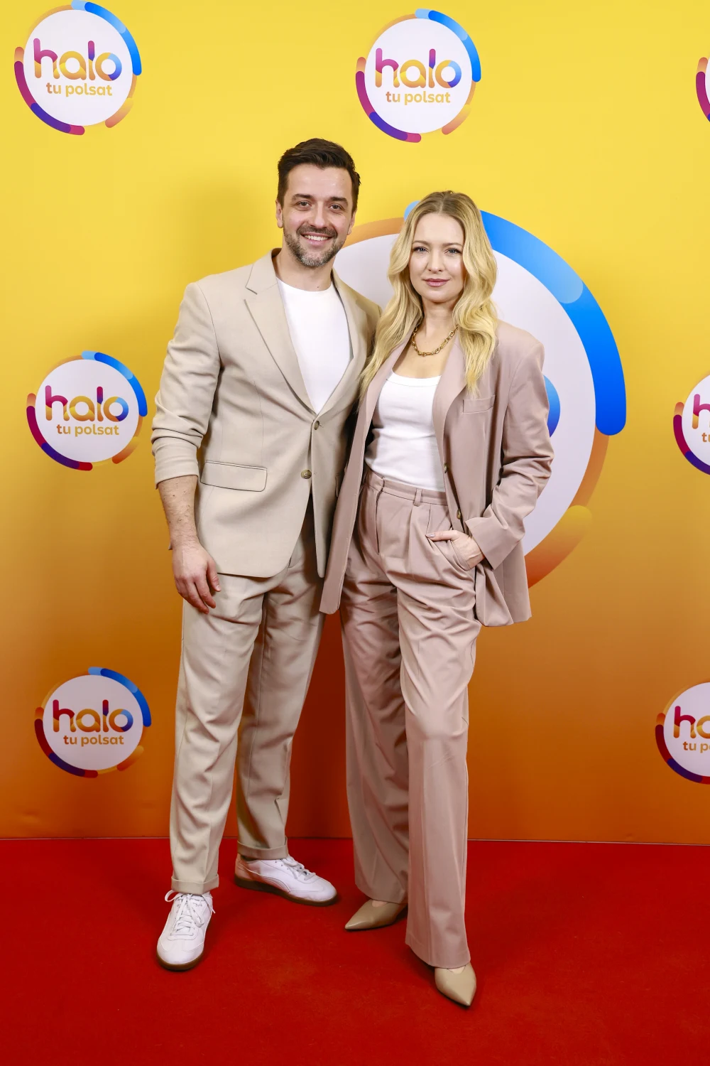 Dwoje uśmiechniętych ludzi ubranych w beżowe garnitury pozuje razem na czerwonym dywanie na tle żółtej ścianki z logo 'halo tu polsat'.