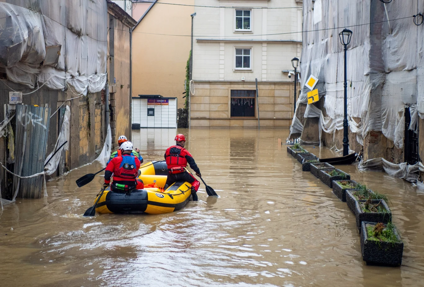 Jedną z największych katastrof naturalnych o szerokim zasięgu jest powódź Trzech ratowników płynie żółtą pontonem ulicą zalaną wodą, otoczoną przez zabezpieczone folią budynki. Poziom wody sięga wysoko, a ratownicy wyposażeni są w kaski i kamizelki.