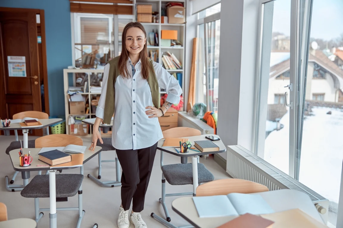 Jak zdobyć przygotowanie pedagogiczne Młoda kobieta stoi w jasnej, nowoczesnej sali lekcyjnej otoczonej pustymi ławkami, książkami oraz przyborami szkolnymi, za nią widać okna z widokiem na zaśnieżone budynki.