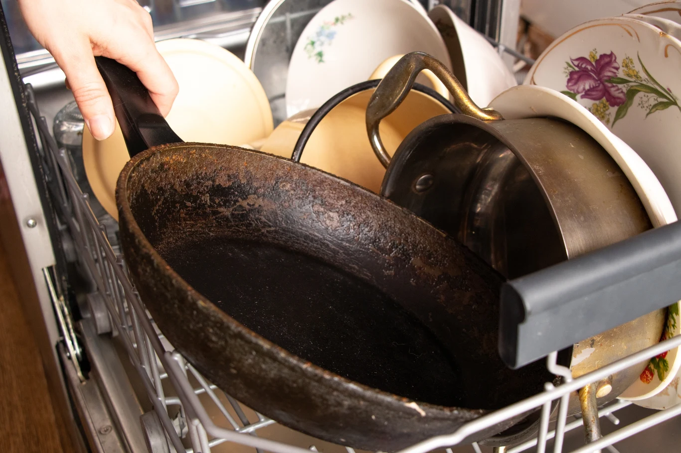 Ręka trzyma rdzewiejącą patelnię umieszczoną w zmywarce wśród talerzy i garnków, widoczne ślady użytkowania naczyń.