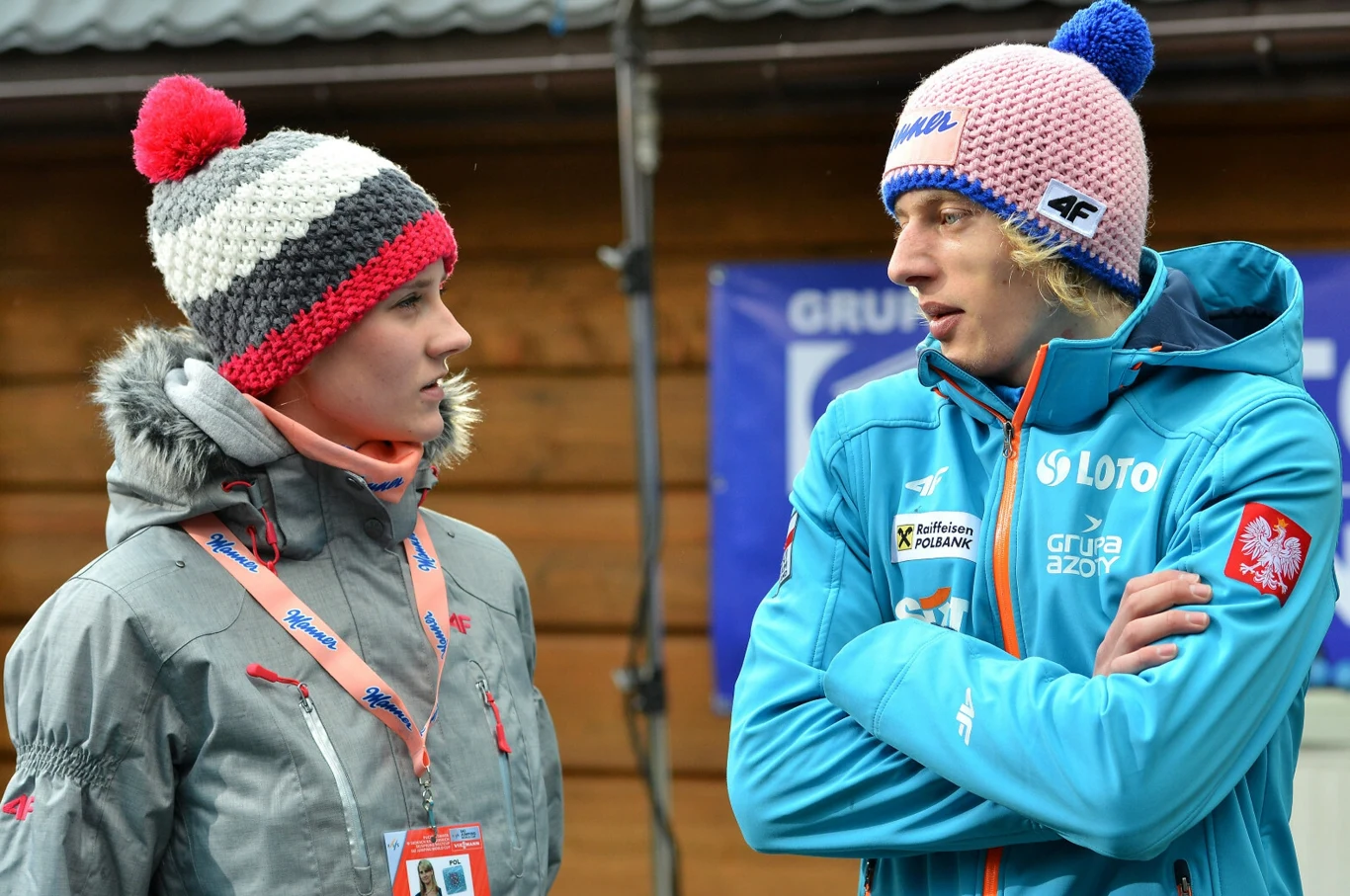 Dwoje młodych ludzi w zimowych czapkach rozmawia, ubrani w sportowe kurtki z widocznymi logotypami sponsorów, w tle drewniana ściana i baner.