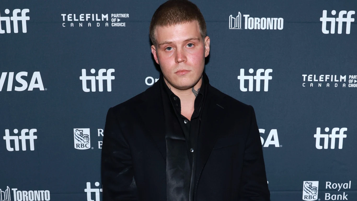 Yung Lean zagra na OFF Festivalu Młody mężczyzna w ciemnej marynarce i czarnej koszuli stoi na tle ścianki z logotypami sponsorów i wydarzenia filmowego, wyglądając poważnie.