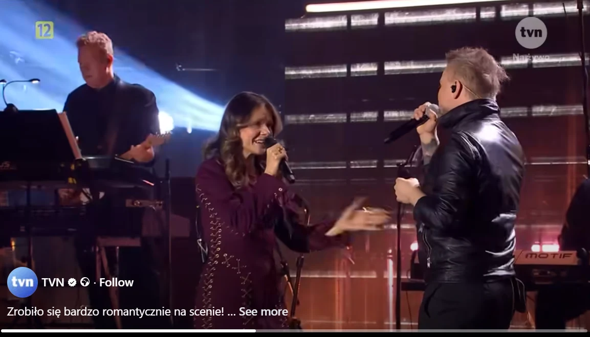 Dwoje wokalistów wykonuje utwór na scenie w otoczeniu muzyków, artystka śpiewa do mikrofonu z uśmiechem, a wokalista zwraca się do niej, za nimi znajduje się klawiszowiec i sceniczne oświetlenie.