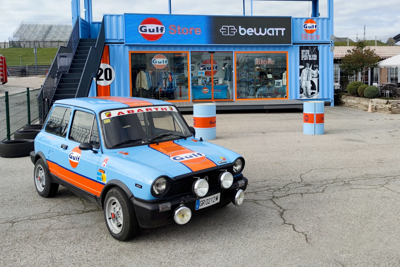 Amerykańska sieć Gulf wchodzi do Polski Niebieski samochód marki Abarth z pomarańczowymi pasami i logotypami Gulf, ustawiony na parkingu przed sklepem motoryzacyjnym Gulf Store, który mieści się w kontenerze. W tle widoczna konstrukcja ze schodami i duże okna sklepu, na których eksponowane s...