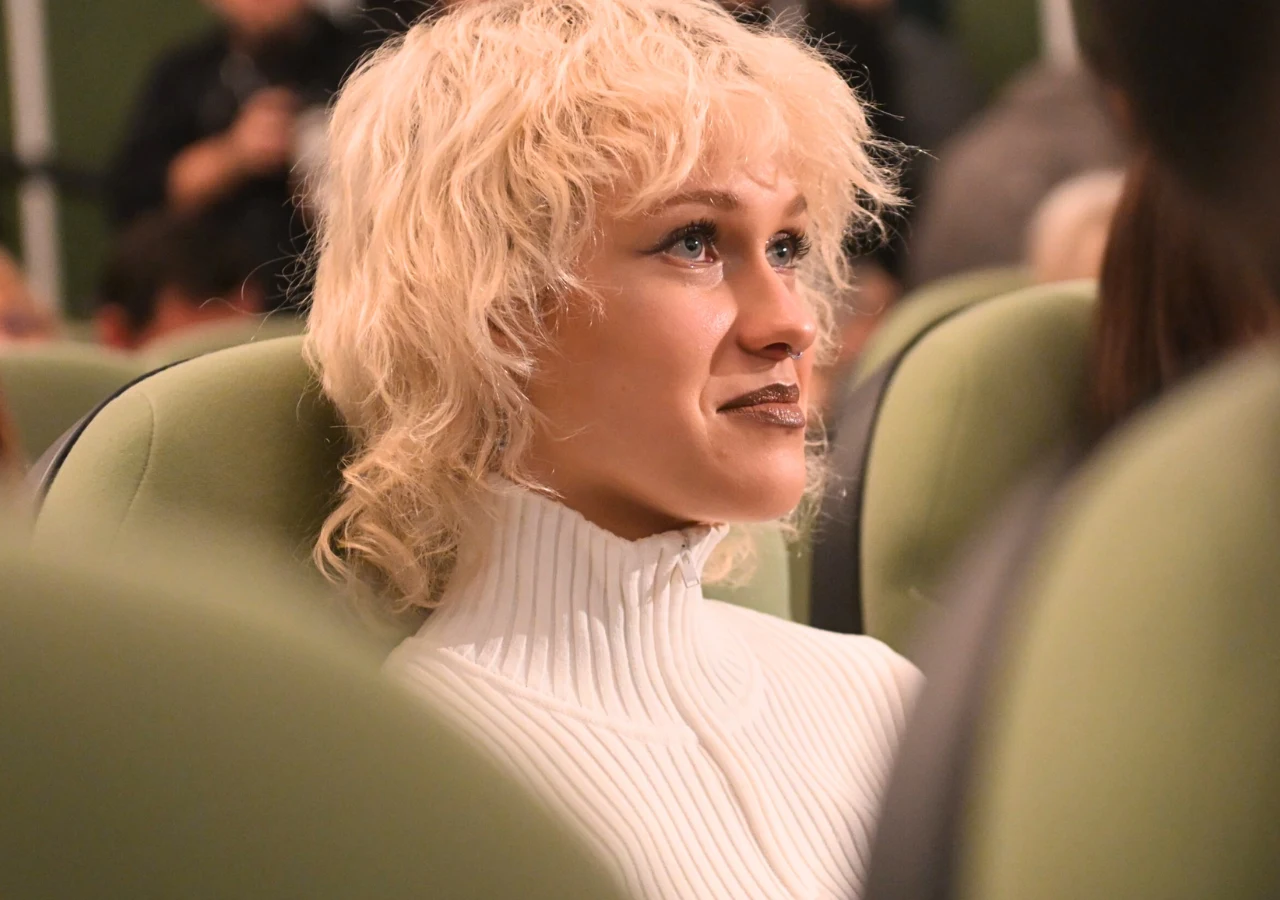 Helena Englert Helena Englert z blond kręconymi włosami siedzi na zielonym krześle w pomieszczeniu pełnym ludzi, ubrana w biały golf, skupiona, patrząca lekko w górę z wyrazem zainteresowania.