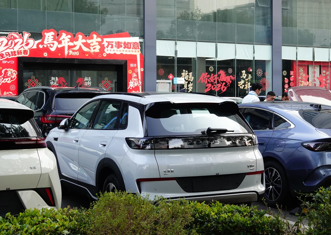 Tanie chińskie e-auta mogą przestać być tanie Nowoczesny biały samochód elektryczny zaparkowany tyłem wśród innych pojazdów na parkingu przed przeszklonym budynkiem ozdobionym czerwonymi dekoracjami i napisami po chińsku.