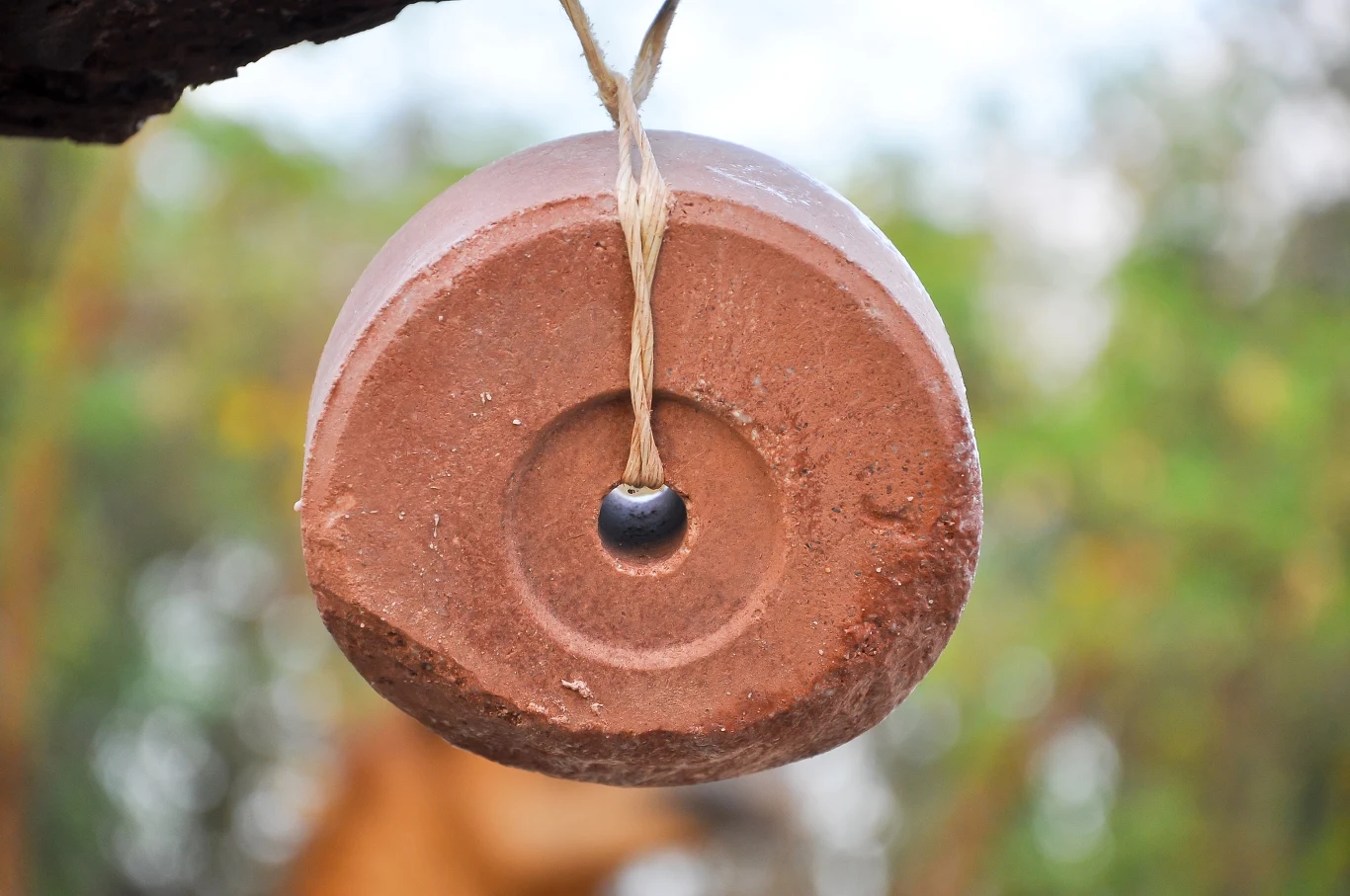 Gliniany dzwonek wiszący na sznurku, w centrum otwór z czarnym koralikiem, w tle rozmazana roślinność.