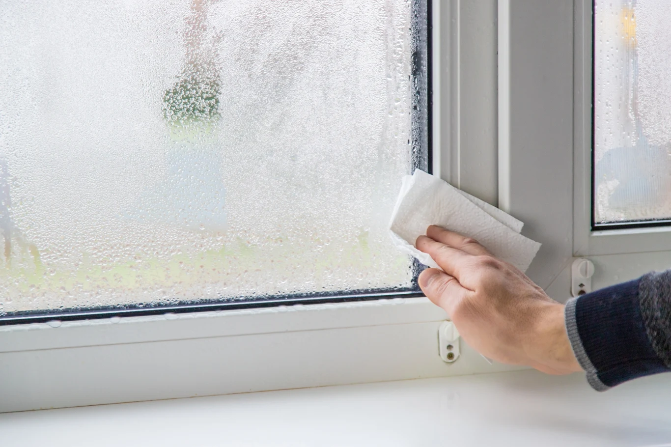 Ręka osoby wyciera zaparowaną szybę okna za pomocą papierowego ręcznika, na ramie okiennej widoczne są krople wody.