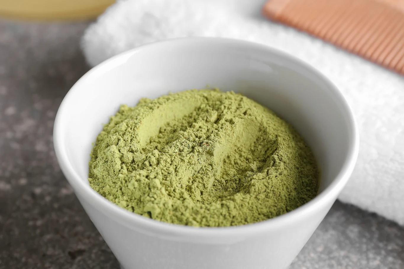 Biała miseczka wypełniona zielonym proszkiem ustawiona na kamiennym blacie, w tle widoczny biały ręcznik i brązowy grzebień.