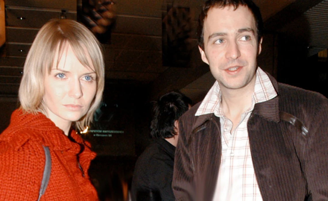 Dwie osoby, kobieta w czerwonym swetrze oraz mężczyzna w jasnej koszuli i ciemnej kurtce, stoją razem w pomieszczeniu o przytłumionym oświetleniu. W tle widoczne rozmyte sylwetki innych osób oraz elementy wystroju wnętrza.