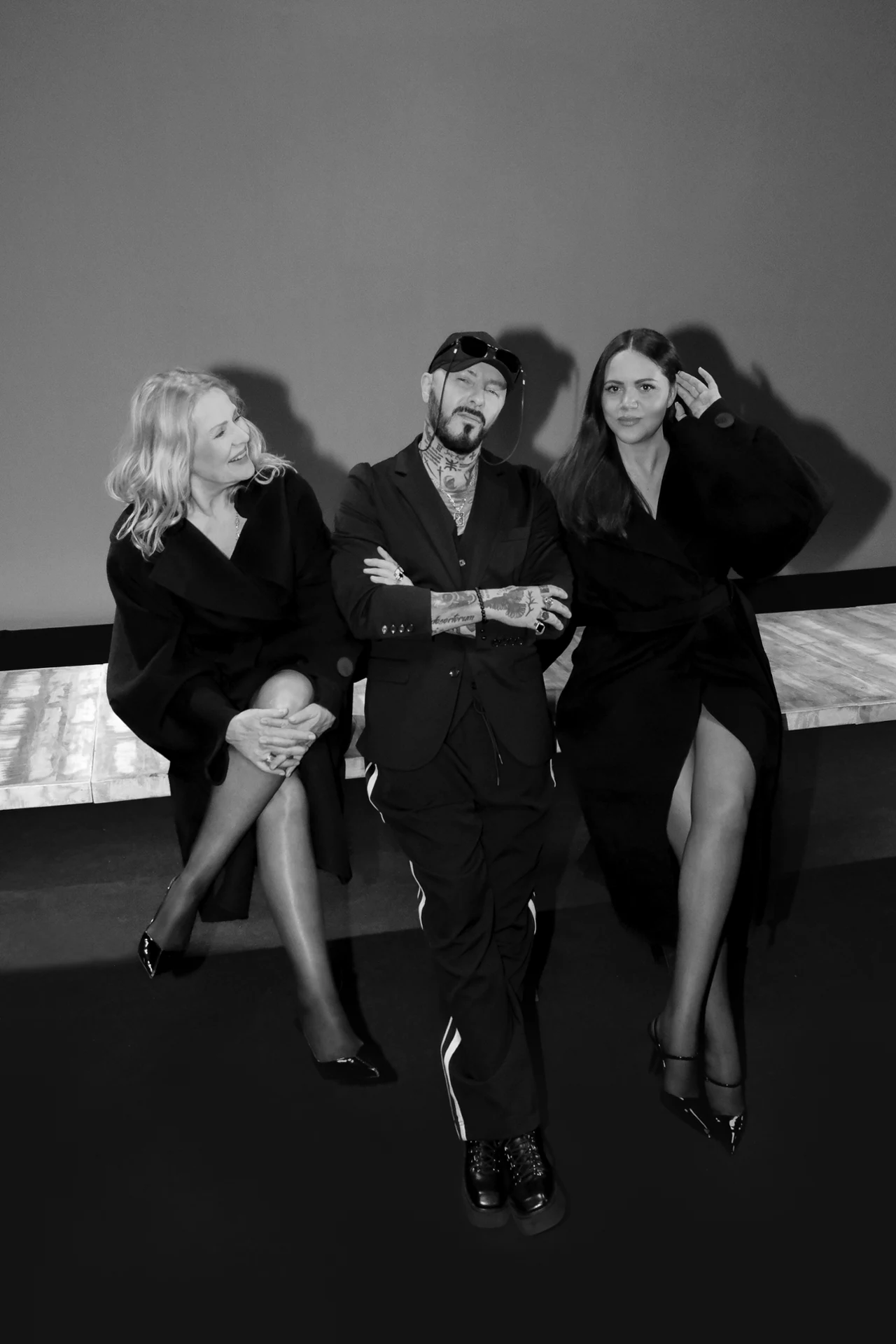 Trzy osoby w eleganckich strojach siedzą obok siebie na tle jasnej ściany, centralnie znajduje się mężczyzna w ciemnym garniturze z tatuażami na rękach, po jego bokach dwie kobiety w czarnych sukienkach z odkrytymi nogami, których ułożenie wskazuje na ...