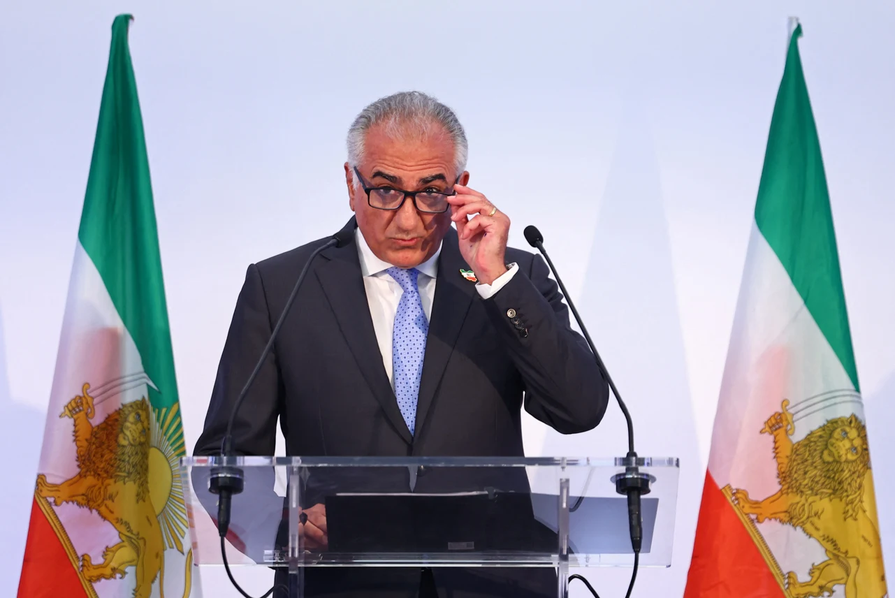 Mężczyzna w garniturze i okularach stoi przy mównicy, poprawiając okulary, w tle widoczne są dwie flagi z symbolem lwa z mieczem.