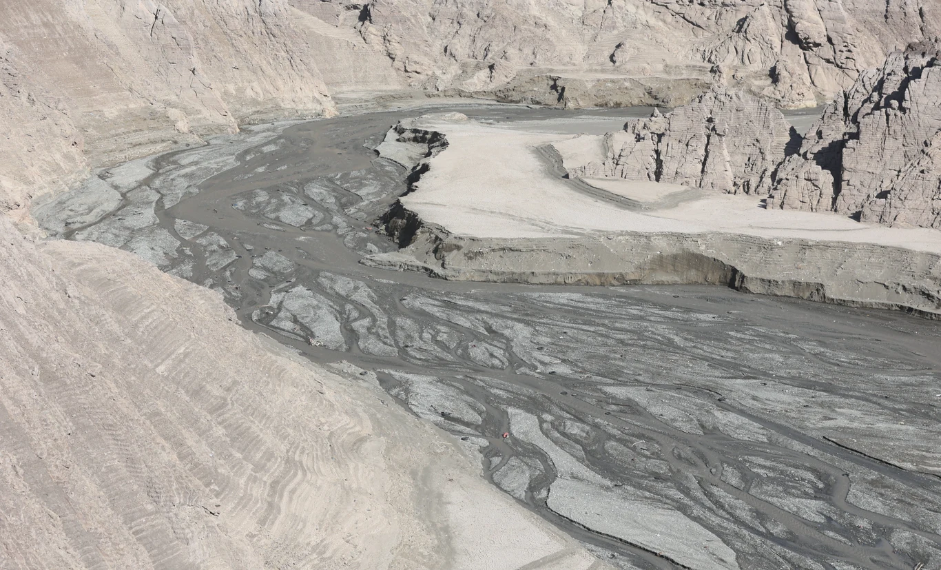 Suchy, jałowy krajobraz z szarym, błotnistym korytem rzeki płynącej między wysokimi, kamienistymi zboczami górskimi; dominują odcienie szarości i beżu, powierzchnia ziemi spękana i pozbawiona roślinności.