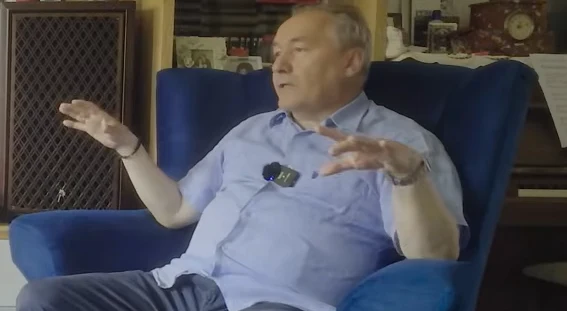 Starszy mężczyzna siedzący w niebieskim fotelu, ubrany w jasnoniebieską koszulę, z mikrofonem przypiętym do ubrania, gestykuluje rękami podczas rozmowy. W tle znajduje się głośnik, zegar i regał z książkami.