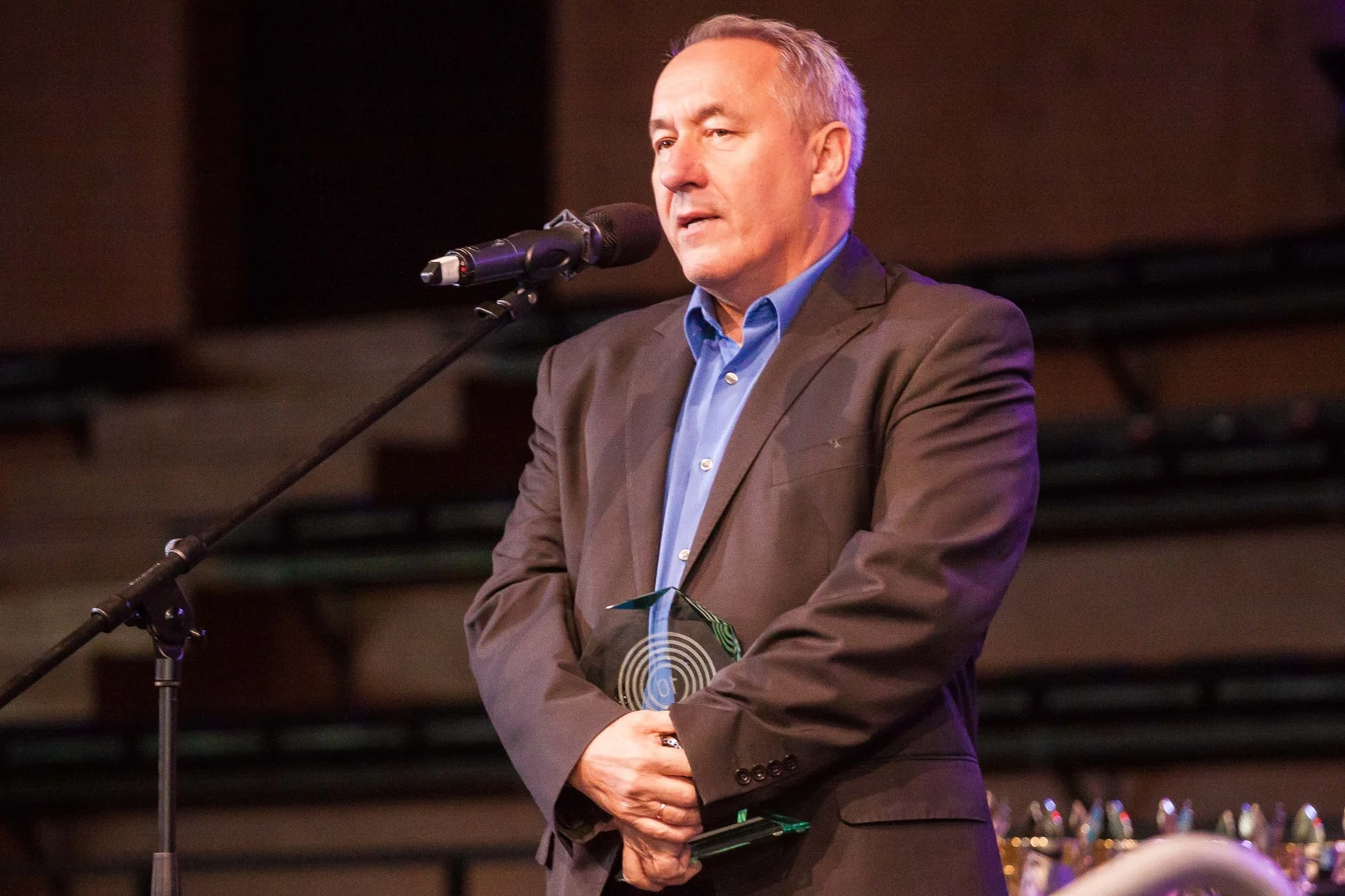 Mężczyzna w garniturze trzymający nagrodę, stojący przy mikrofonie na scenie podczas uroczystości wręczenia wyróżnień.