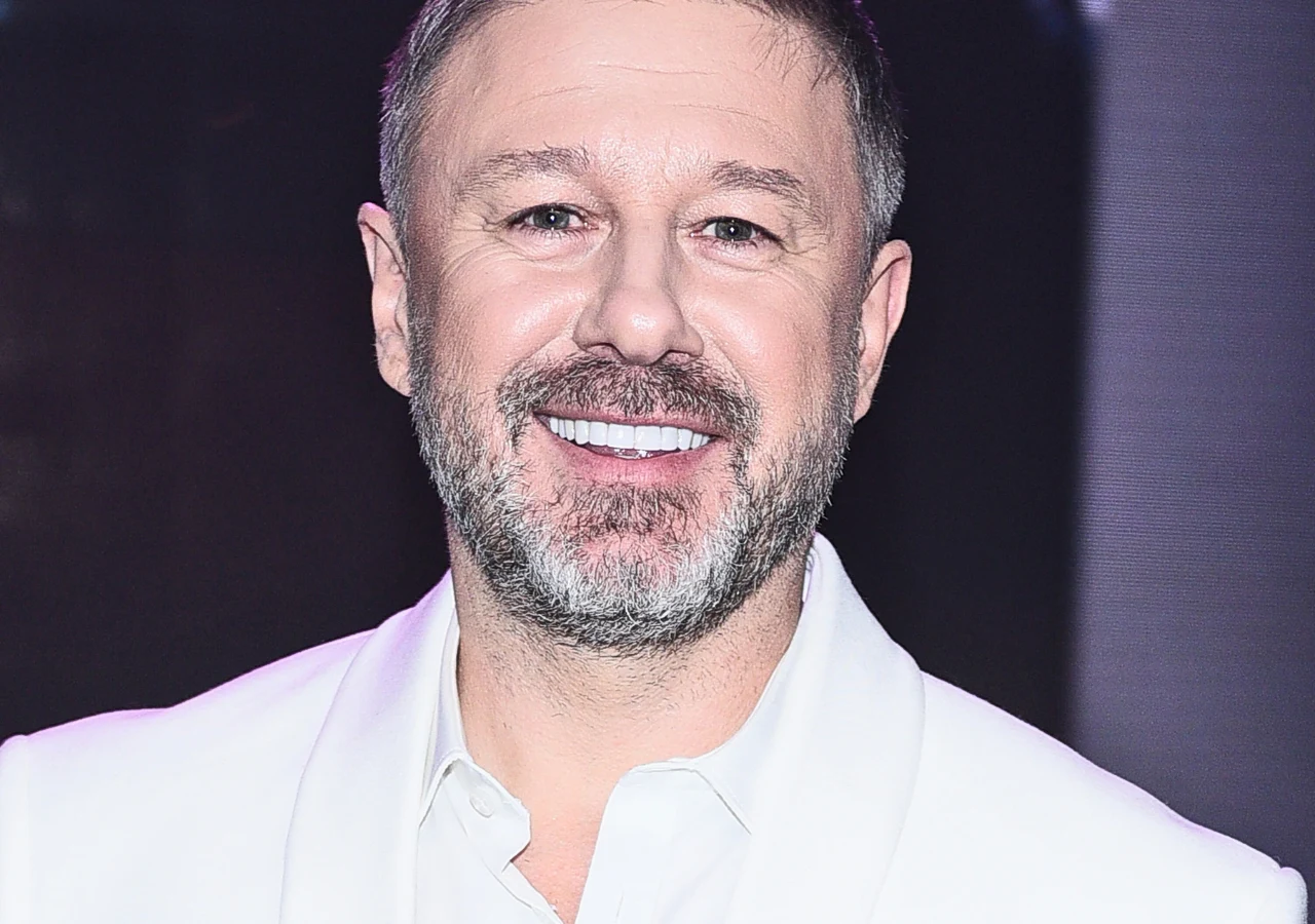 Andrzej Piaseczny Andrzej Piaseczny - Uśmiechnięty mężczyzna z krótkimi włosami i brodą, ubrany w białą marynarkę i koszulę, stoi na ciemnym tle. Jego twarz oświetlona jest z przodu, podkreślając przyjazny wyraz twarzy.