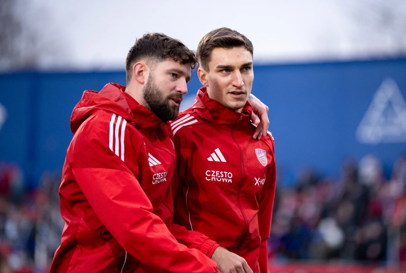 Dwóch mężczyzn w czerwonych sportowych kurtkach z logo drużyny stoi obok siebie na tle rozmytego stadionu, jeden z nich obejmuje drugiego ramieniem, obaj skupieni, otoczeni sportową atmosferą.