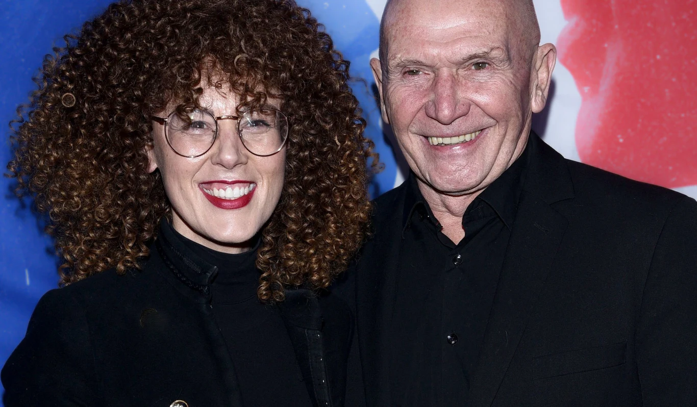 Dwoje uśmiechniętych ludzi pozujących razem, kobieta z kręconymi włosami i okularami ubrana na czarno, mężczyzna łysy w czarnym garniturze, w tle kolorowa grafika.