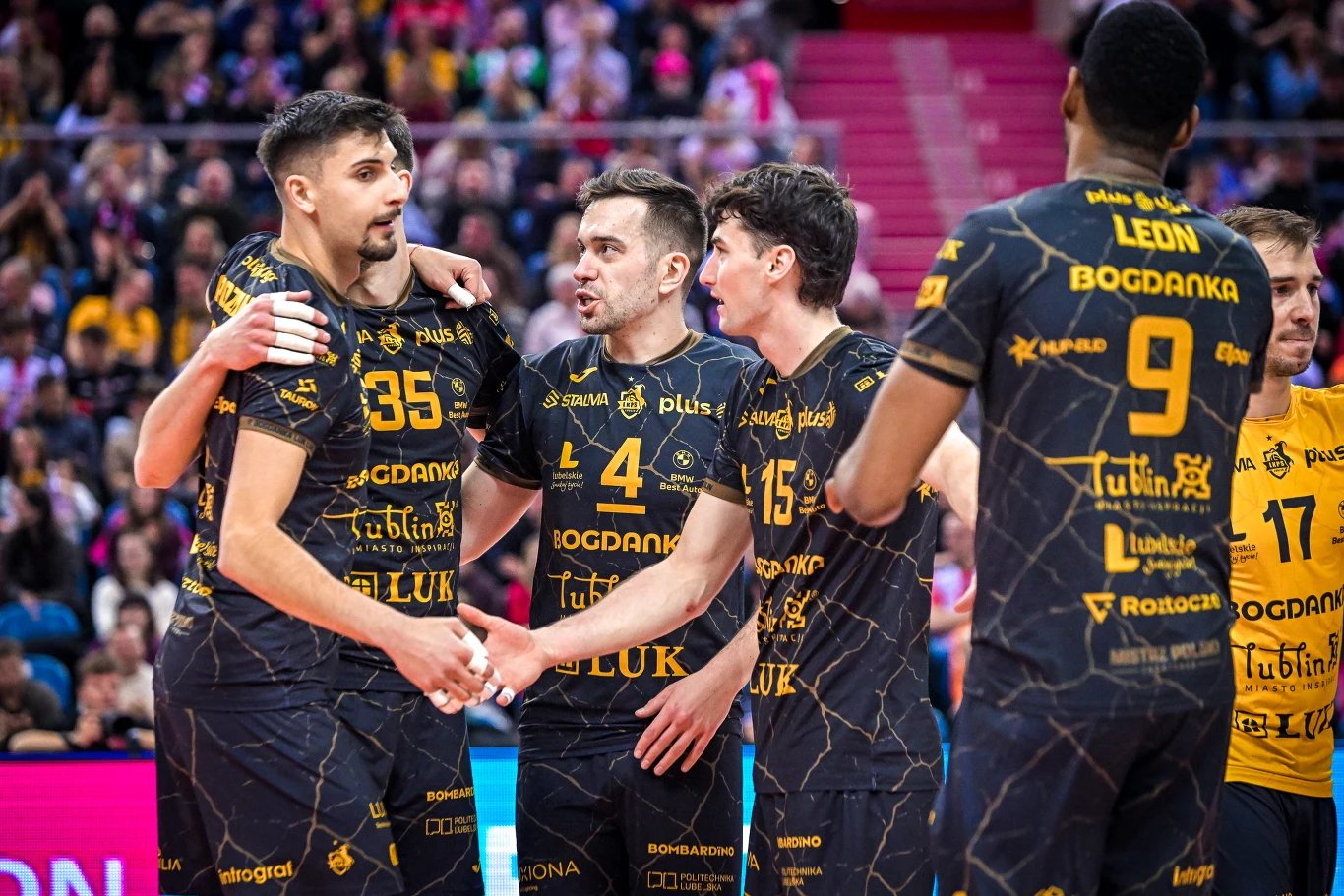 Grupa siatkarzy ubranych w ciemne stroje z żółtymi napisami, stojących razem na boisku podczas przerwy w meczu, otoczona przez widownię na trybunach. Na koszulkach widoczne są nazwiska oraz nazwy sponsorów.