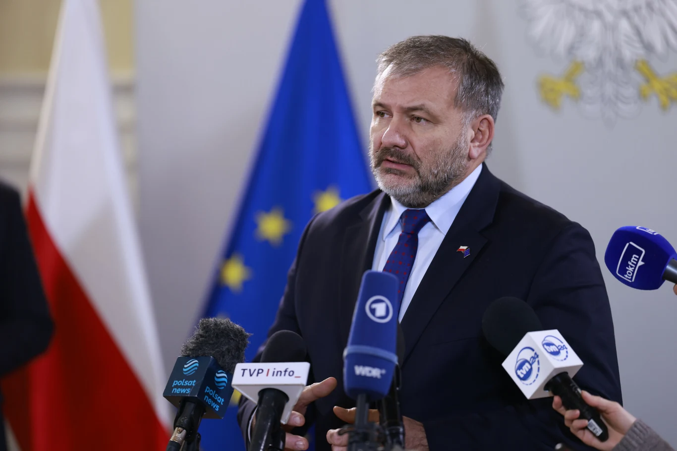 Mężczyzna w garniturze przemawia podczas konferencji prasowej, trzymając kilka mikrofonów należących do różnych stacji telewizyjnych. W tle widoczne flagi Polski oraz Unii Europejskiej.