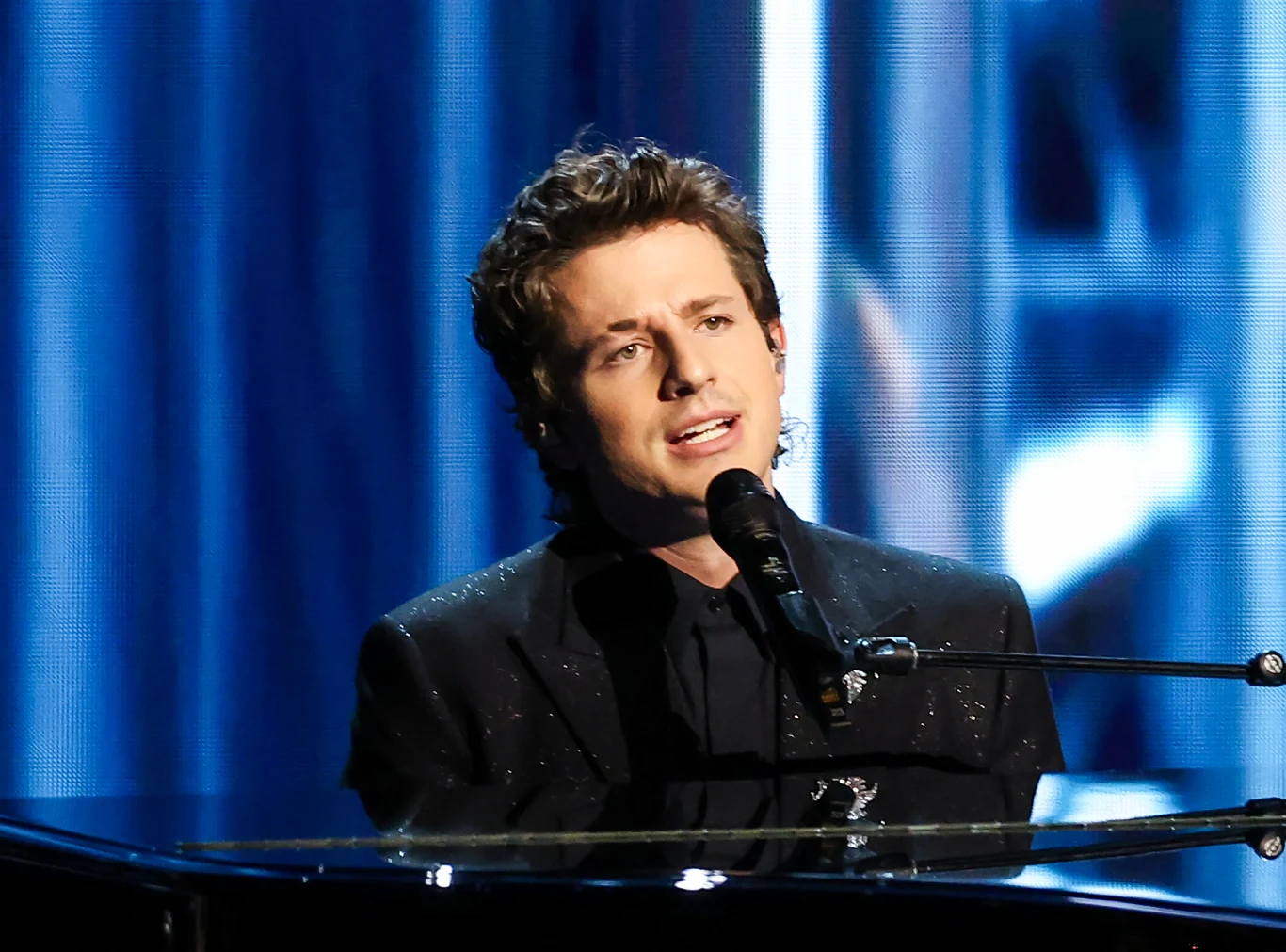 Charlie Puth wystąpi w Warszawie Młody mężczyzna ubrany w elegancki, czarny garnitur gra na fortepianie i śpiewa do mikrofonu, a w tle widoczne są jasne światła i niebieskie reflektory.