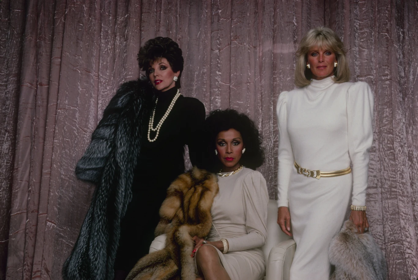 Trzy elegancko ubrane kobiety w stylowych kreacjach z lat 80., ubrane w futra oraz perły, pozują na tle różowej, błyszczącej kotary.