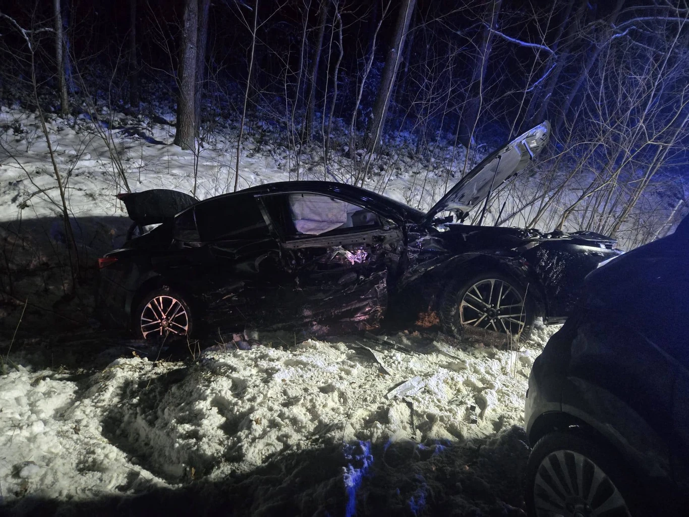 Czarny samochód osobowy znajdujący się w rowie przy leśnej drodze, uszkodzony po wypadku, z otwartą maską oraz widocznymi śladami zniszczeń, wokół śnieg i drzewa.