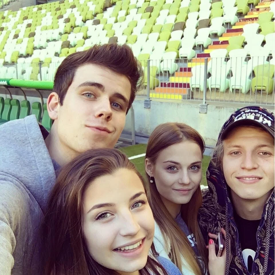 Czwórka młodych ludzi robi selfie na tle pustych trybun stadionowych, uśmiechnięci, w swobodnych ubraniach, w jasnym świetle dziennym.