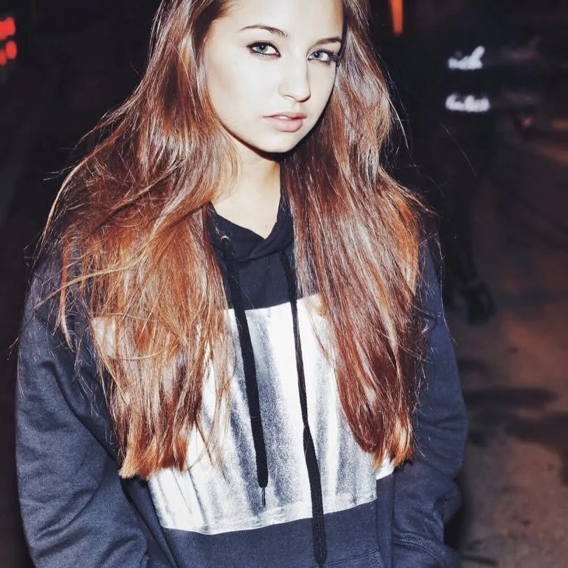 Młoda kobieta z długimi, brązowymi włosami ubrana w czarną bluzę z jasnym prostokątnym wzorem, stoi na tle rozmytego nocnego miasta, patrząc bezpośrednio w obiektyw.