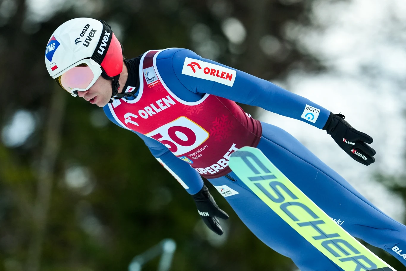Zawodnik w niebieskim kombinezonie i czerwonej kamizelce startowej podczas skoku narciarskiego w locie, w tle rozmyte drzewa, na kasku widoczne logo sponsora.