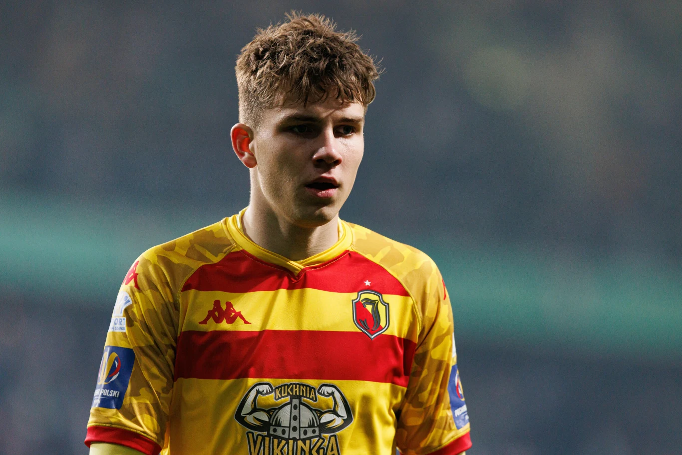 Młody piłkarz w żółto-czerwonej koszulce drużyny Jagiellonia Białystok stoi na stadionie podczas meczu, z lekko rozchylonymi ustami i skoncentrowaną miną.