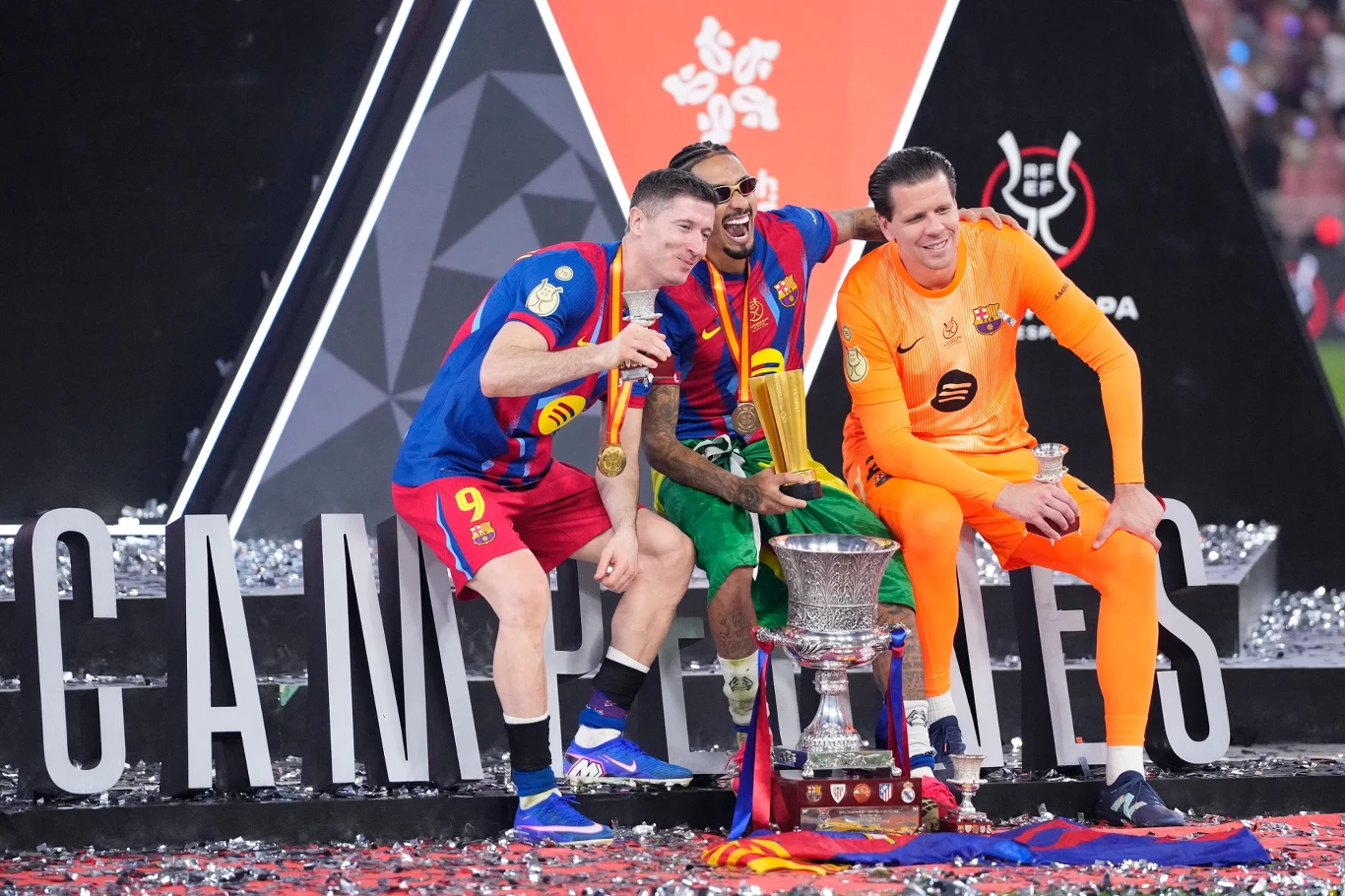 Trzech piłkarzy klubu FC Barcelona w kolorowych strojach drużynowych siedzi obok siebie na scenie świętując zdobycie trofeum, w tle duże litery oraz dekoracje związane z wygraną, wokół konfetti.