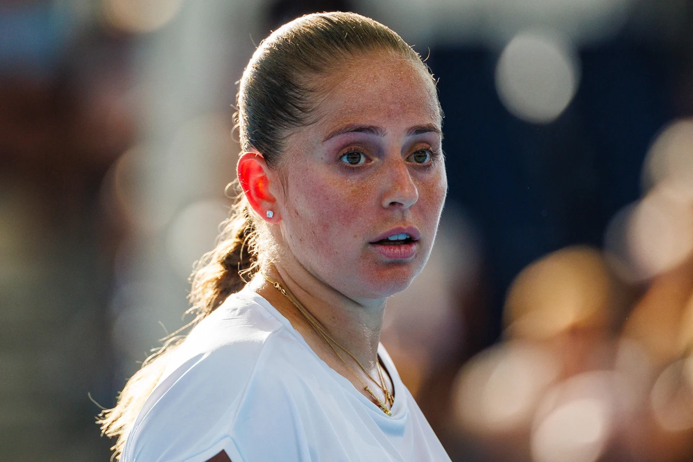 Młoda kobieta z jasnobrązowymi włosami spiętymi w kucyk, ubrana w biały sportowy strój, stoi zwrócona bokiem, patrząc z powagą przed siebie. Tło jest rozmyte, podkreślając główną postać.