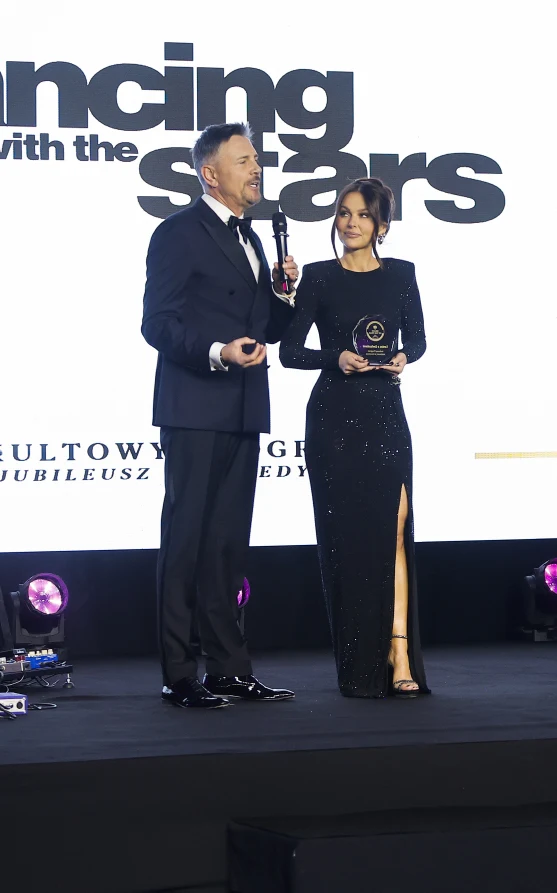 Dwóch elegancko ubranych prowadzących na scenie podczas gali programu rozrywkowego, mężczyzna trzyma mikrofon i mówi, kobieta w długiej, czarnej sukni z rozcięciem trzyma w dłoniach nagrodę. W tle widoczny jasny ekran z logo programu Dancing with the S...