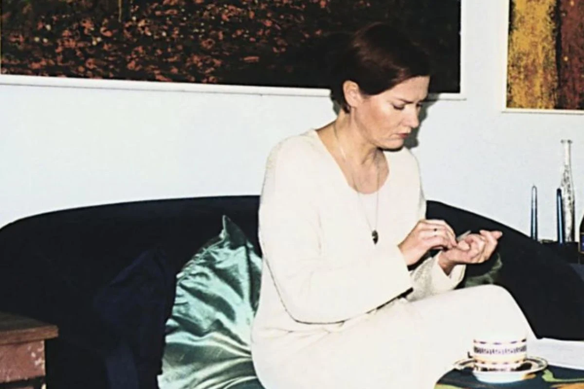 Kobieta w jasnym ubraniu siedzi na ciemnej kanapie z poduszką, skupiona na własnych dłoniach, obok filiżanka na małym stoliku i dekoracje ścienne w tle.