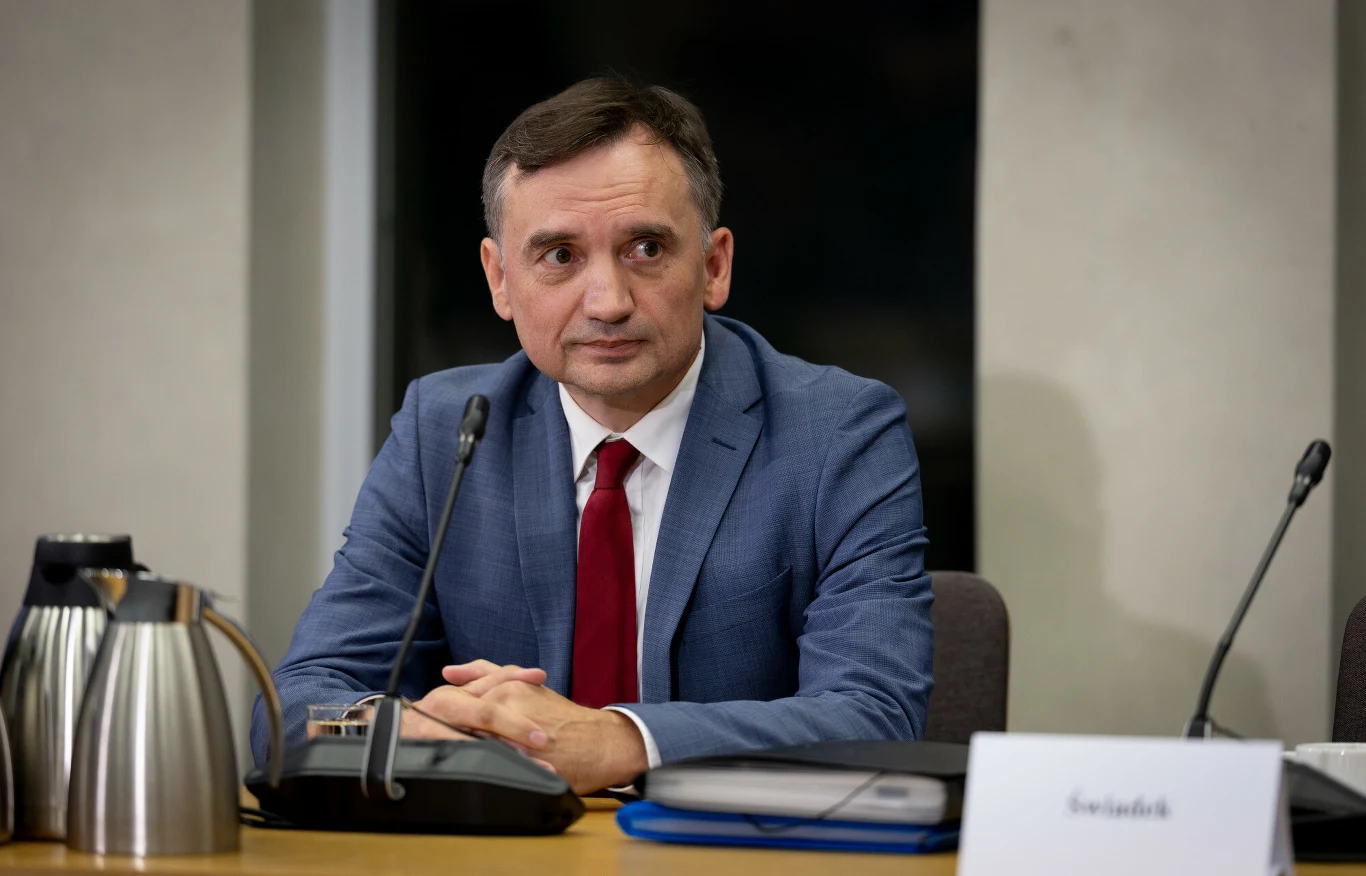 Zbigniew Ziobro odniósł się do kwestii azylu otrzymanego na Węgrzech Mężczyzna w garniturze i czerwonym krawacie siedzi za stołem konferencyjnym, przed nim zestaw do kawy, mikrofon oraz dokumenty. W tle jasna ściana i czarne okno.